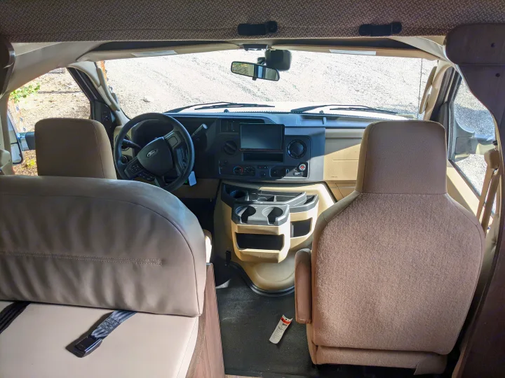 RV rental interior view: forward into the driver's cockpit with steering wheel, dashboard, front seats, and windshield beyond.. Winnebago Minnie Winnie 2022