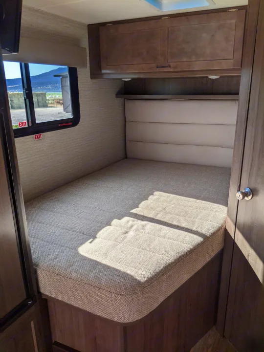 RV rental bedroom: a cozy interior with a comfortable bed, overhead cabinets, and sunlit window overlooking a rural scene.. Winnebago Minnie Winnie 2022