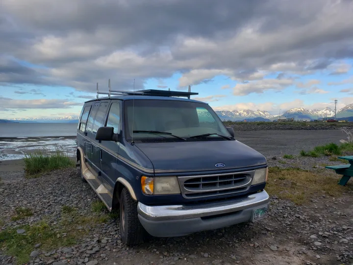 A blue Ford van, perfect for an RV rental adventure, parked on an Alaskan rocky beach facing snow-capped mountains.. Ford Econoline Club Wagon 1998
