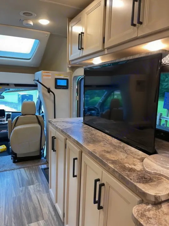 Modern RV rental interior: light cabinets, a patterned countertop, and a TV, with the driver's area visible.. Thor Gemini 2021
