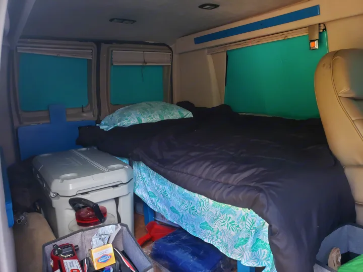 RV rental interior: a comfy bed with tropical sheets, teal window covers, and a large cooler.. Ford Econoline Club Wagon 1998