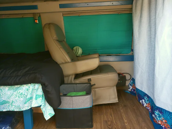 Inside an RV rental, featuring a cozy bed, reclining chair, small fan, and electrical power unit, with teal window insulation.. Ford Econoline Club Wagon 1998