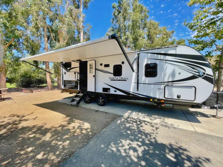 A white North Trail travel trailer RV rental, with an awning out, is parked at a sunny campsite.. Heartland North Trail 2021
