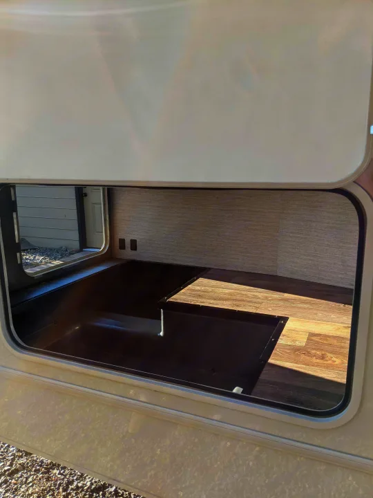 An RV rental's open exterior storage compartment reveals a deep well, wood floor, and power outlets. Gravel is visible outside.. Winnebago Minnie Winnie 2022