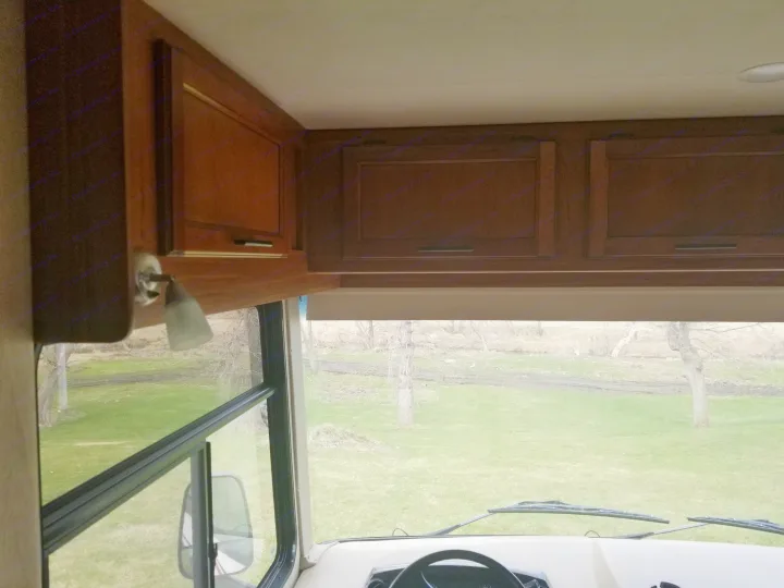 Inside an RV rental, showing overhead wooden cabinets, dashboard, and the view through the windshield of a grassy field with trees.. Jayco Alante 2016