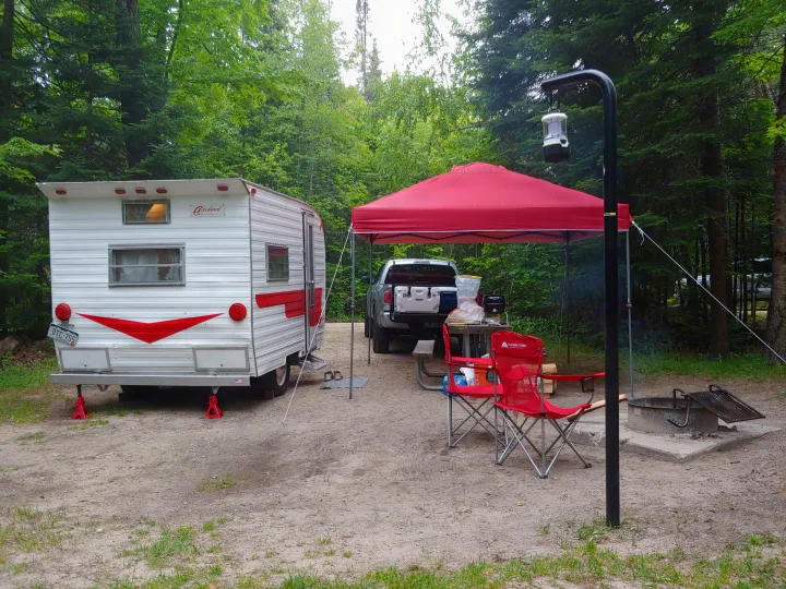 An outdoor camping scene features a vintage rv rental travel trailer, a pickup truck, red canopy, and fire pit at a wooded campsite.. Adams Cardinal Travel Trailer 1972