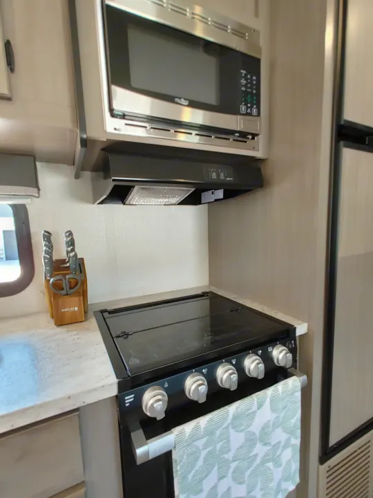 View of a compact RV rental kitchen featuring a microwave, range hood, stove, and knife block on the counter.. Starcraft Travel Trailer 2021