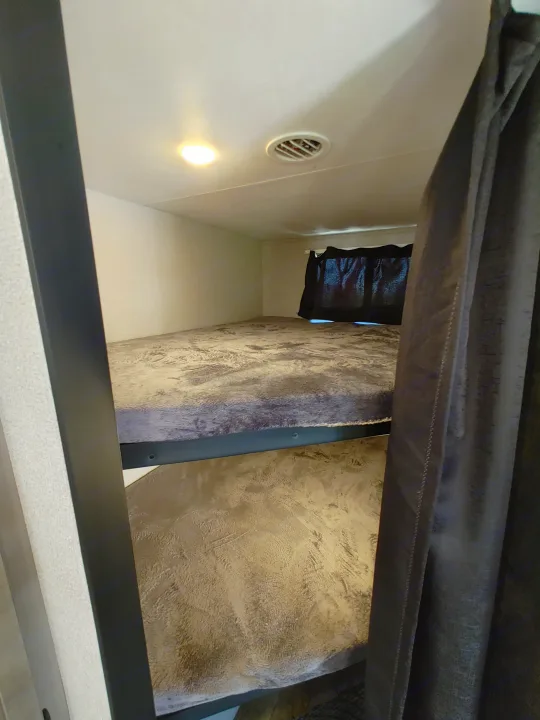 RV rental bunk beds feature patterned bedding, a window curtain, ceiling light, and vent, offering cozy sleeping space.. Starcraft Travel Trailer 2021