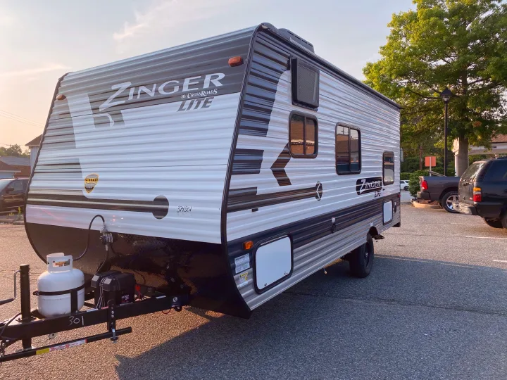 New Zinger Lite travel trailer, ideal for an rv rental, parked outdoors. Features include a propane tank and battery on the hitch.. Crossroads Zinger 2021