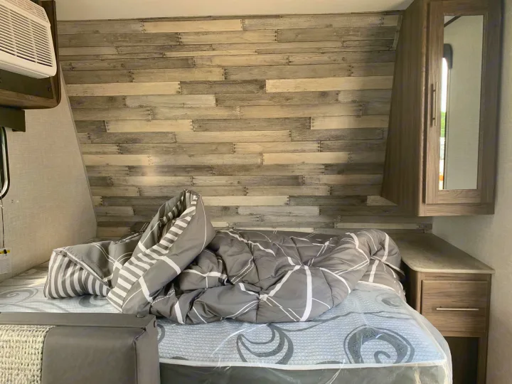 Inside an RV rental: a compact bedroom with a bed, grey comforter, wood-look wall, AC unit, and nightstand.. Crossroads Zinger 2021