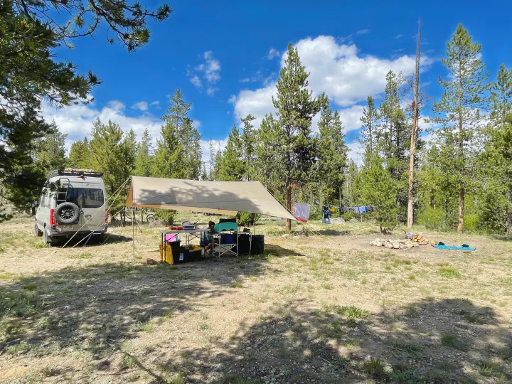An RV rental is perfectly set up at a serene forest campsite. A large awning shelters an outdoor kitchen and chairs, with laundry drying nearby under blue skies.. Mercedes-Benz Sprinter 2020