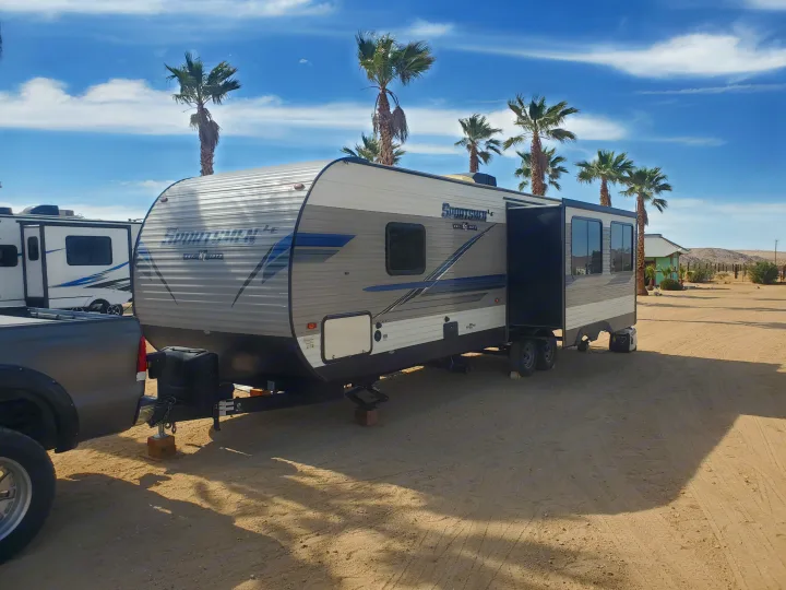 An RV rental travel trailer is parked in a sandy desert campground, surrounded by palm trees under a clear blue sky.. K-Z Manufacturing Sportsmen 2019