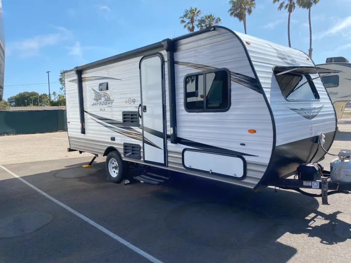 This white and gray Jay Flight SLX travel trailer RV rental is parked on an asphalt lot under a sunny sky with palm trees, ready for travel.. Jayco Jay Flight 2019