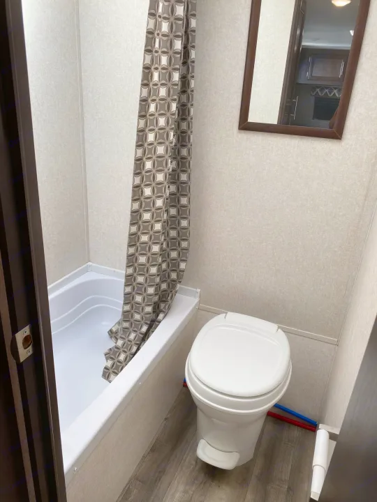 Compact RV rental bathroom featuring a shower/tub combo with patterned curtain, a toilet, and a framed mirror.. Jayco Jay Flight 2019