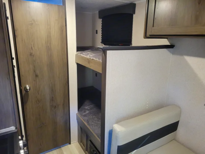 Compact RV rental interior showing bunk beds with gray bedding, a light sofa, and a wooden door. Ideal for family travel.. Coachmen Clipper 2021