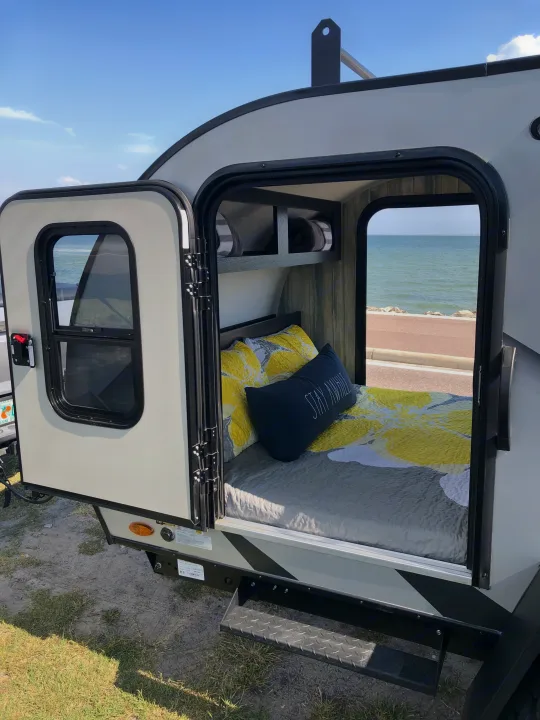 Cozy rv rental teardrop camper bedroom with 'Stay Awhile' pillow, open to ocean views and coastal road.. Braxton Creek Free Solo 2021
