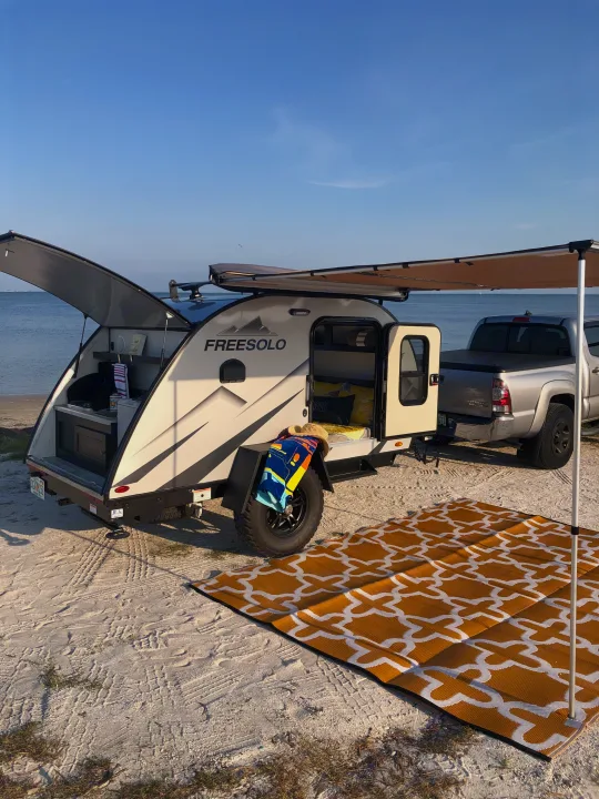 A Freesolo teardrop RV rental is perfectly set up for beach camping. A truck is attached, with an awning and outdoor rug by the ocean.. Braxton Creek Free Solo 2021