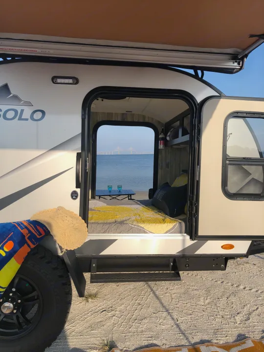 RV rental: Camper on sandy shore, open door reveals cozy bed, table, and stunning bridge view over water.. Braxton Creek Free Solo 2021