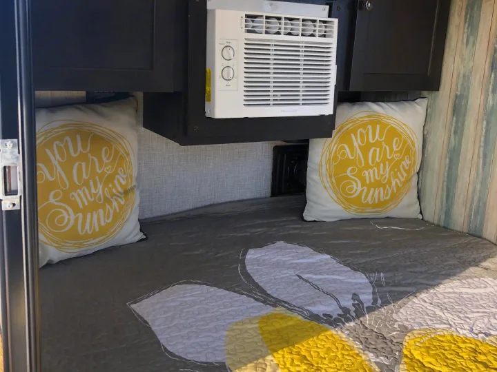 Cozy RV rental bedroom features a gray & yellow bed, "you are my sunshine" pillows, and an AC unit for comfort.. Braxton Creek Free Solo 2021