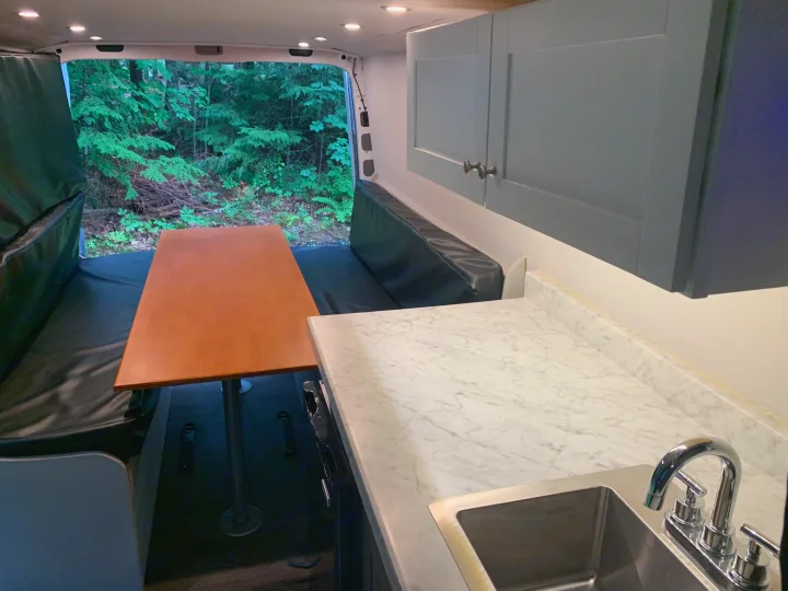 RV rental interior featuring a kitchen with sink, dining area with table & benches, and a forest view from the back.. Mercedes-Benz B class campervan MB 140 2014