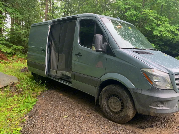 Dark green camper van, likely an rv rental, with its side door open on a gravel path in a lush forest.. Mercedes-Benz B class campervan MB 140 2014