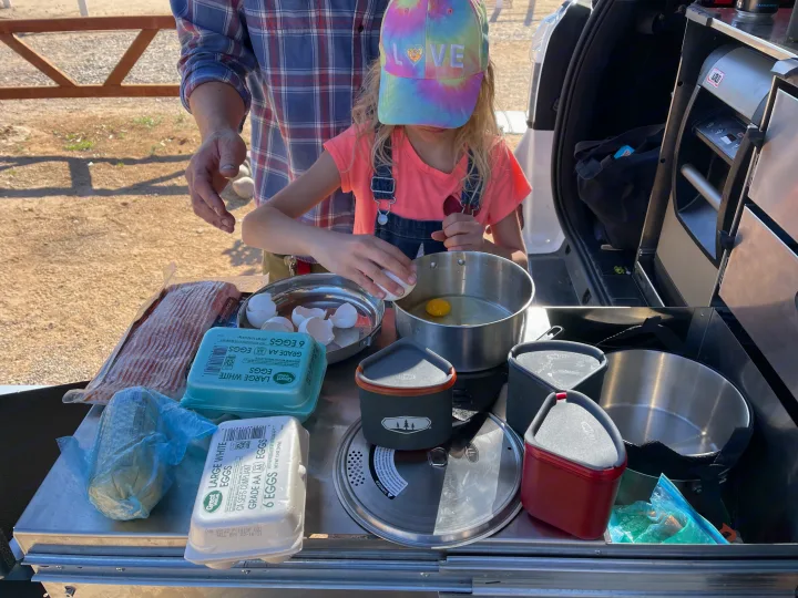 Making breakfast outdoors from an RV rental: a child cracks eggs with an adult assisting at a vehicle's camping kitchen.. Toyota 4Runner 2021