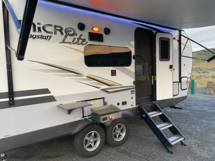 An rv rental travel trailer parked with its awning extended and lights on. Features include an outdoor griddle and entry steps.. Flagstaff Micro Lite 2021