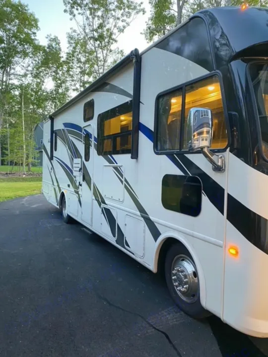 A modern white RV motorhome with black/blue stripes, parked. Interior lights glow, perfect for your next rv rental journey.. Thor Motor Coach A.C.E 2021