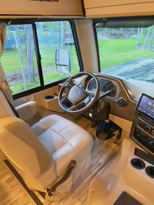 Inside an RV rental, the driver's cockpit features a light seat and dashboard, with trees and green landscape visible outside.. Thor Motor Coach A.C.E 2021