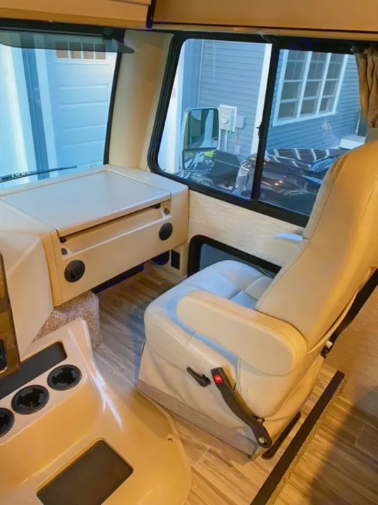 RV rental driver's area: light-colored seats, dashboard with cup holders, house exterior visible through windows.. Thor Motor Coach A.C.E 2021