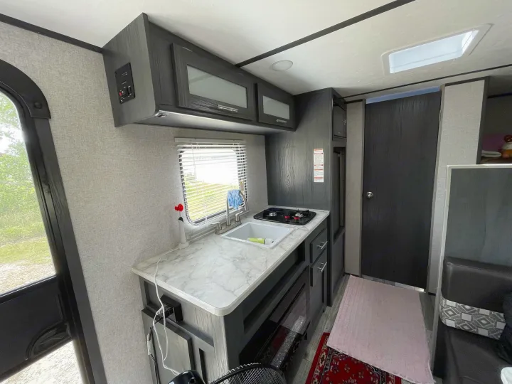 Inside an RV rental: a compact, modern kitchen with sink, stove, and overhead cabinets, alongside a small living space with seating.. Dutchmen Aspen Trail 2021