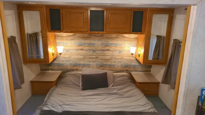 Cozy RV rental bedroom featuring a comfortable bed, overhead wooden cabinets, ambient lighting, and wood-paneled wall.. Dutchmen Lite 2003