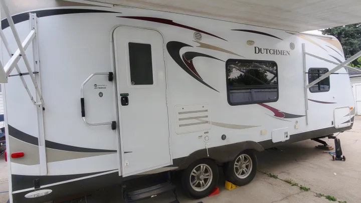 White RV rental travel trailer parked outdoors. Features include an extended awning, main door, windows, and multiple wheels.. Dutchmen Dutchmen 2012