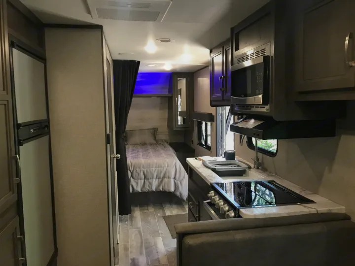 Modern RV rental interior showing a kitchen with sink and stovetop, refrigerator, and a cozy bedroom area.. Jayco qbw 2021