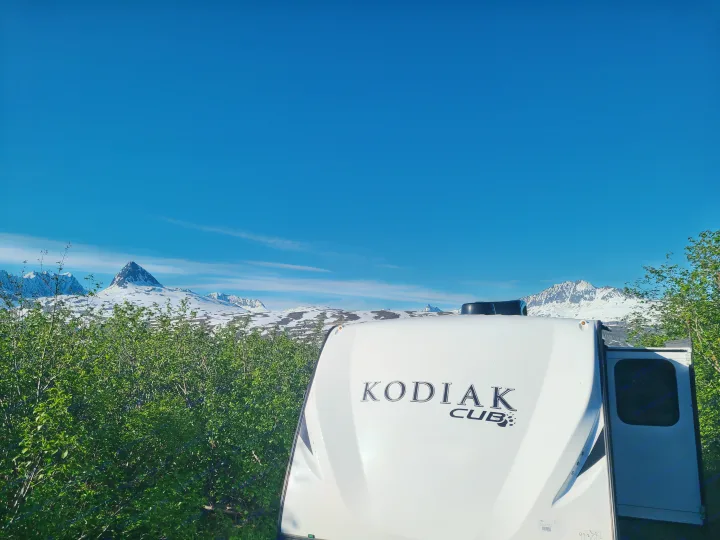 RV rental adventure: A white Kodiak Cub camper overlooks stunning snow-capped mountains and green bushes under a clear blue sky.. Dutchmen Kodiak 2021