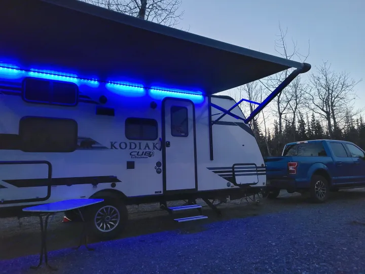 Night camping with an RV rental: Kodiak Cub travel trailer, illuminated by blue awning lights, hitched to a blue truck.. Dutchmen Kodiak 2021