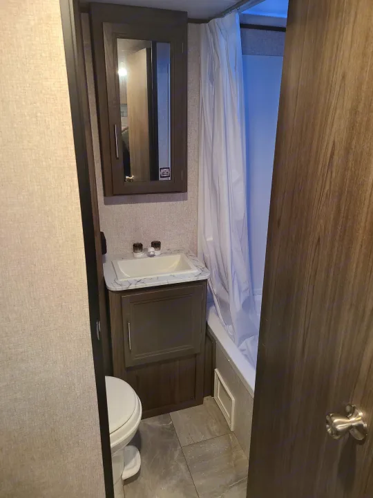 Clean RV rental bathroom showcasing a compact toilet, vanity with sink, mirror cabinet, and shower with curtain.. Dutchmen Kodiak 2021