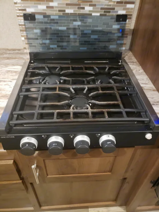 A three-burner Greystone RV cooktop with cast iron grates, control knobs, and a reflective glass cover, common in an RV rental.. Shasta Oasis 2019
