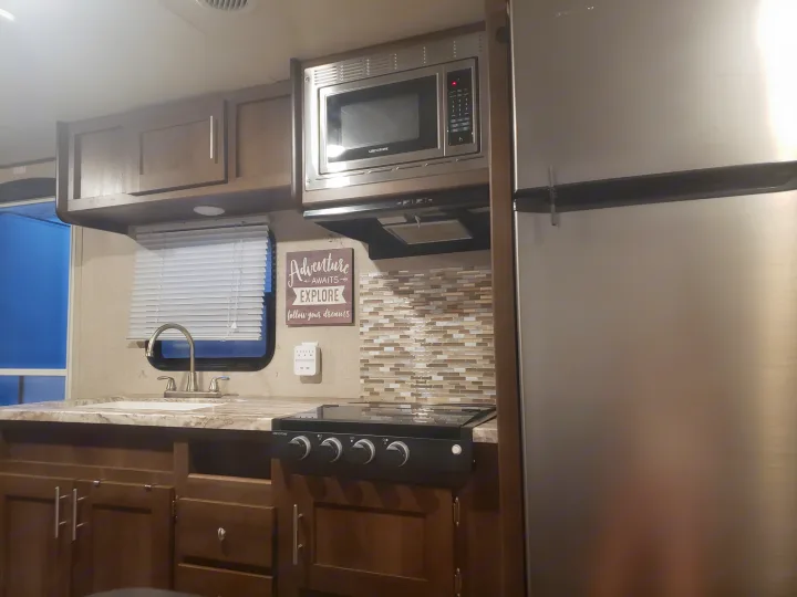 RV rental kitchen interior: stainless steel microwave, refrigerator, stovetop, sink, and wood cabinets. A decorative "Adventure" sign hangs on the wall.. Shasta Oasis 2019