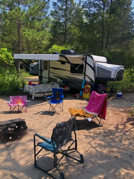 This serene campsite features a hybrid travel trailer, perfect for an RV rental getaway. Chairs and a fire pit invite outdoor relaxation.. Starcraft Launch 2018