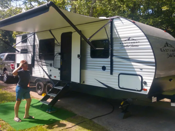 A large, modern RV rental with an extended awning is set up at a sunny campsite, with a person standing by its steps.. East to West Della Terra 2021