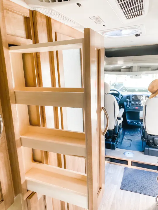 Inside this **RV rental**: a light wood pull-out pantry, partially open, with the driver's cabin visible beyond. Smart camper van storage.. Coachmen Nova 2021