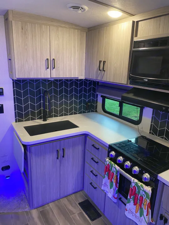 Explore this RV rental kitchen: modern light wood cabinets, black chevron backsplash, appliances, & ambient blue lighting.. Keystone Passport 2021