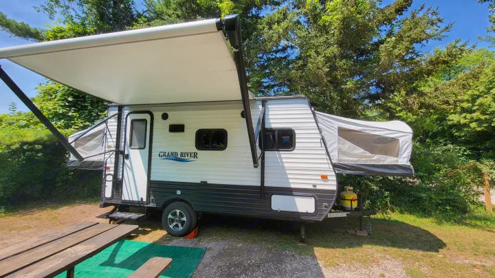 This RV rental is a white hybrid travel trailer with an extended awning, parked at a sunny campsite with a picnic table.. Forest River 16RB 2019