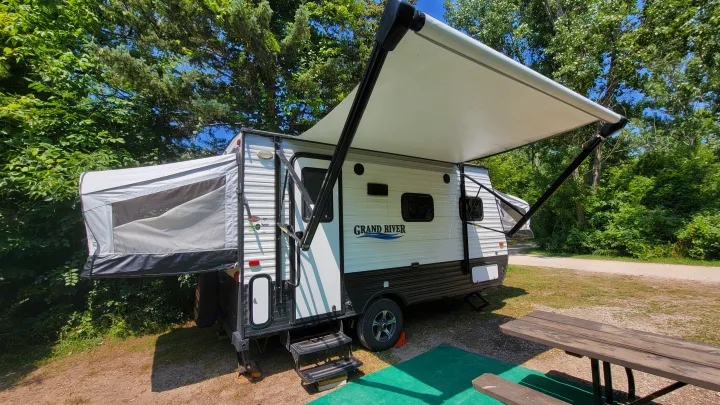 This RV rental, a Grand River travel trailer with an extended awning and pop-out, is perfectly set up at a shady campsite.. Forest River 16RB 2019