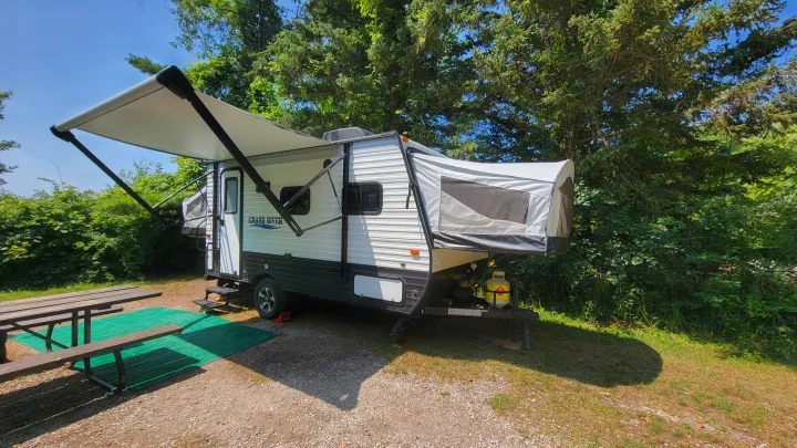 A Grand River RV rental, a travel trailer with extended awning, parked at a sunny campsite. Features a picnic table and green mat.. Forest River 16RB 2019