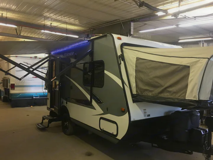 This grey and white hybrid travel trailer, perfect for your next RV rental, is parked indoors with its awning extended and blue lights on.. Jayco Jay Feather 2016