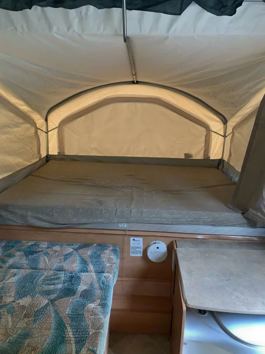 Inside a pop-up RV rental, featuring a spacious bed, light fabric walls, and a small built-in counter area.. Coachmen Sport 2007