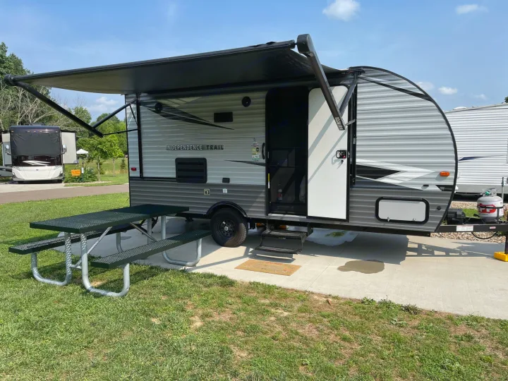RV rental travel trailer set up at a sunny campsite with an extended awning & picnic table.. Forest River Independence Trail 2021
