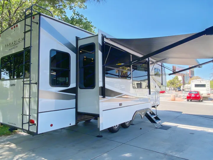 White RV rental with extended slide-outs, a deployed awning, and a rear ladder, set up at a sunny camp area.. Alliance RV Paradigm 2021